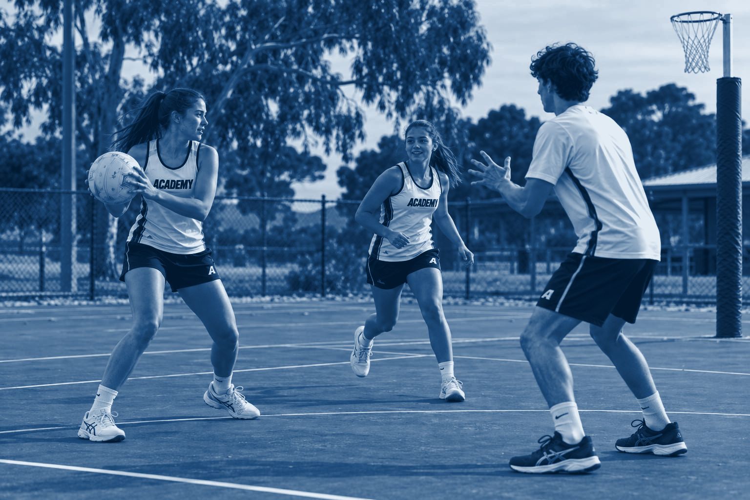 Three Academy players practise a passing drill on a tree-lined outdoor netball court